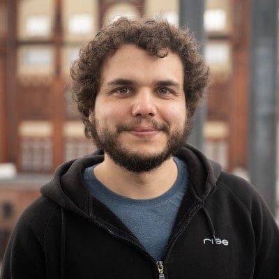 a closeup portrait of a man with curly dark hair and a beard wearing a black zipup hoodie over a blue tshirt he smiles gently offering a warm expression the background features a blurred building with large windows hinting at an urban setting digital production A close-up portrait of a man with curly dark hair and a beard, wearing a black zip-up hoodie over a blue t-shirt. He smiles gently, offering a warm expression. The background features a blurred building with large windows, hinting at an urban setting.