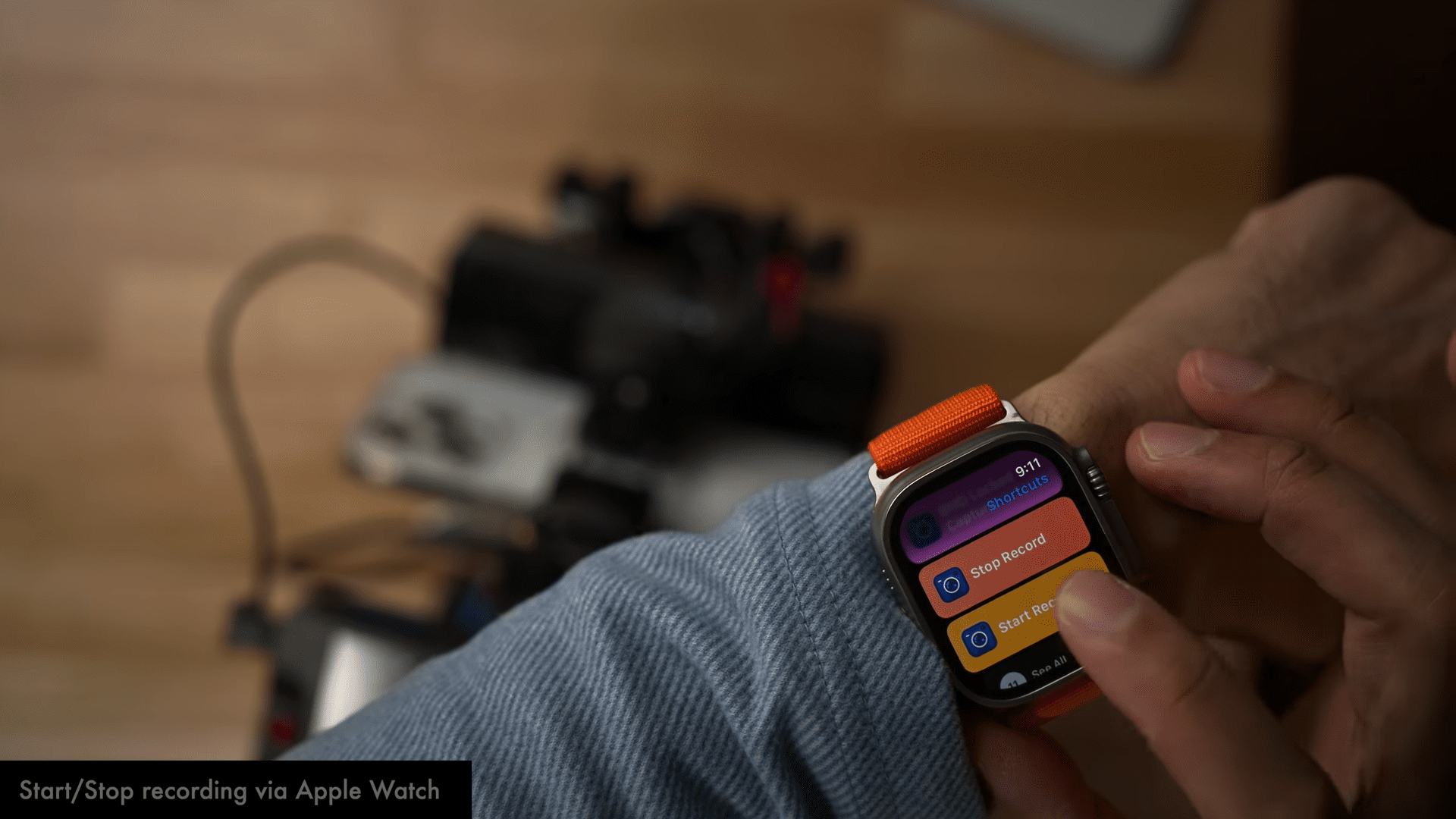 a closeup view of a persons wrist wearing an apple watch with an orange strap the watch screen displays vibrant buttons labeled start research and take photo in the blurred background a piece of equipment is visible with wires coiling over a smooth wooden surface digital production A close-up view of a person's wrist wearing an Apple Watch with an orange strap. The watch screen displays vibrant buttons labeled 'Start Research' and 'Take Photo.' In the blurred background, a piece of equipment is visible, with wires coiling over a smooth wooden surface.