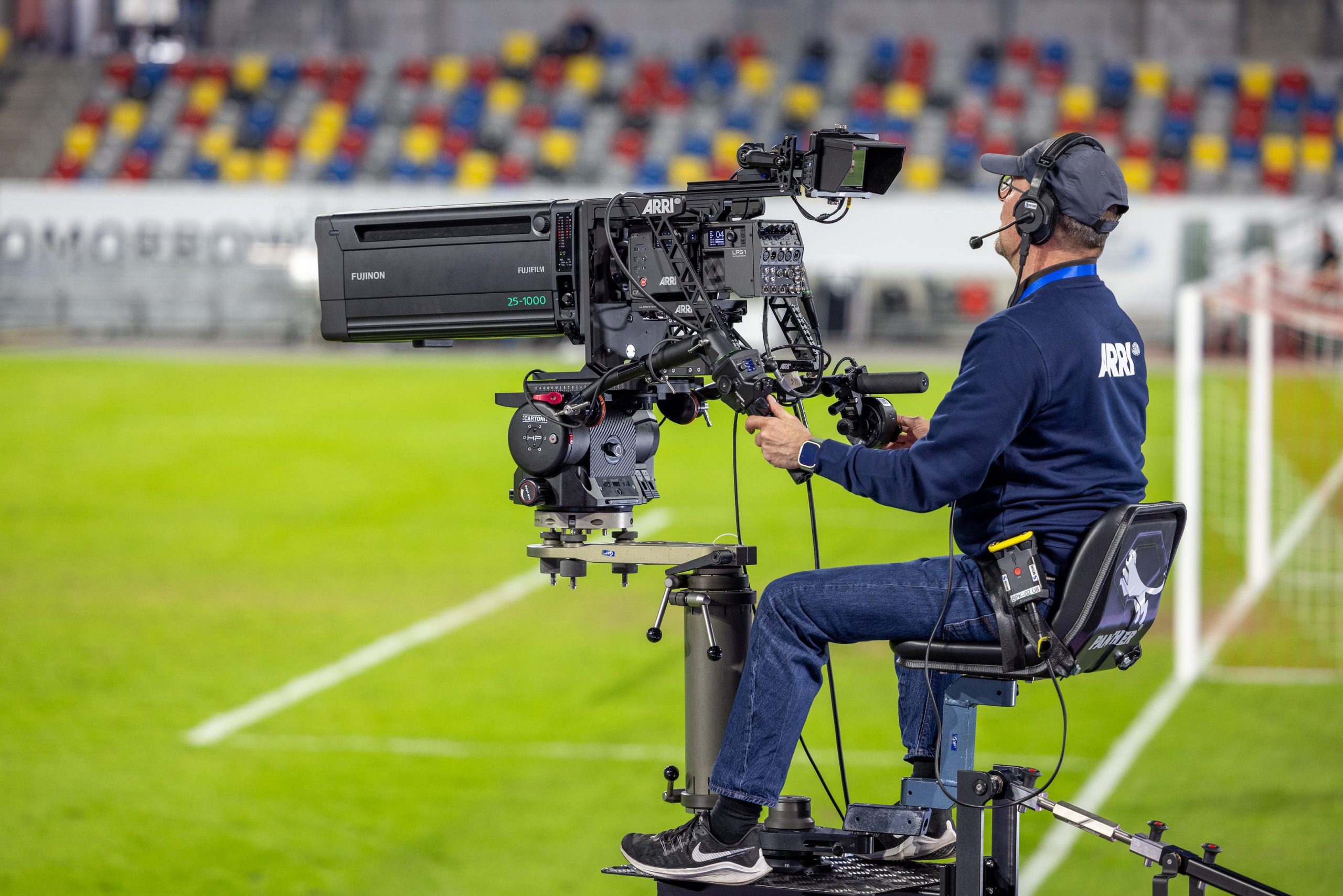 a cameraman operates a professional filming camera mounted on a tall stable tripod he wears headphones and a blue shirt with an insignia focused on capturing the action on the lively football field set against a backdrop of colorful stadium seats digital production A cameraman operates a professional filming camera mounted on a tall, stable tripod. He wears headphones and a blue shirt with an insignia, focused on capturing the action on the lively football field set against a backdrop of colorful stadium seats.