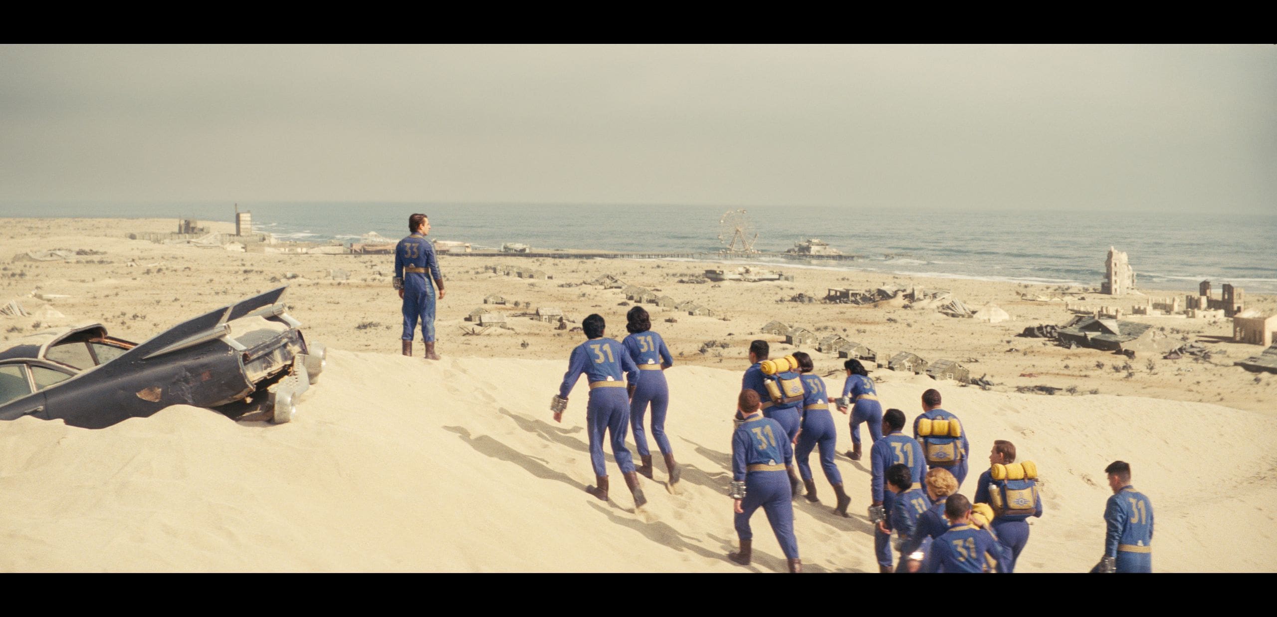 a group of individuals in blue suits trek up a sandy dune overlooking a desolate landscape in the background a sunbleached car lies partially buried in sand while remnants of a lost civilization including ruins and ancient structures scatter along the coastline against a muted sky digital production A group of individuals in blue suits trek up a sandy dune, overlooking a desolate landscape. In the background, a sun-bleached car lies partially buried in sand, while remnants of a lost civilization, including ruins and ancient structures, scatter along the coastline against a muted sky.