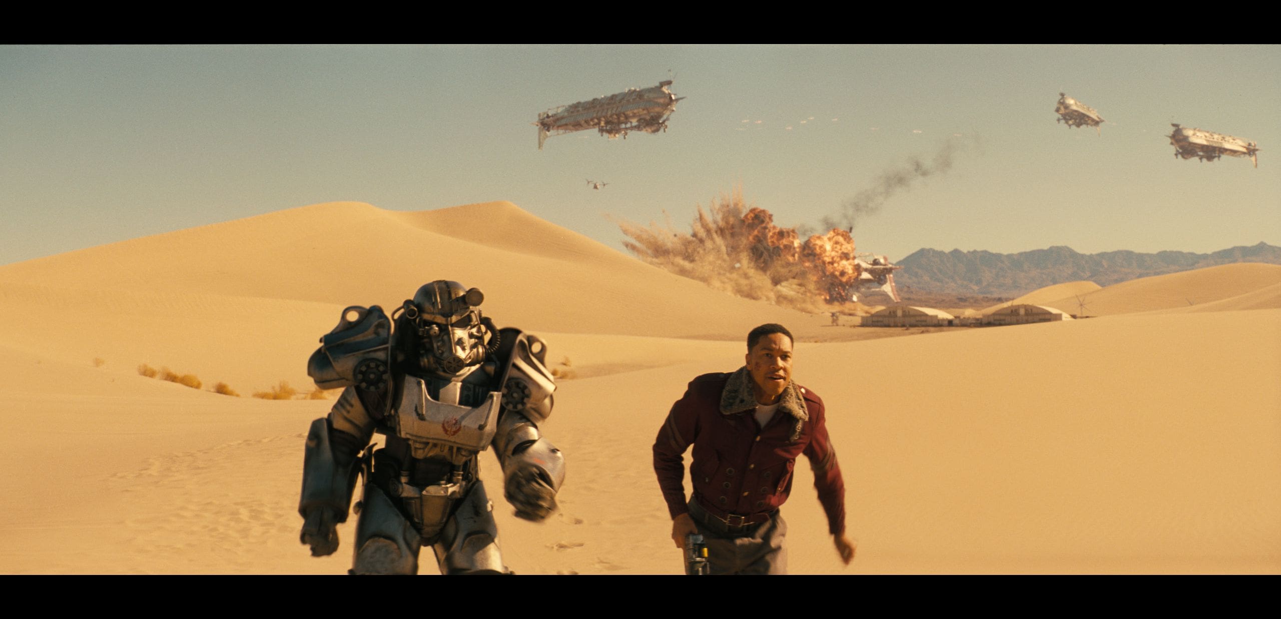 in a vast desert landscape a figure in a futuristic armored suit strides alongside a man in a maroon shirt in the background multiple massive airborne vehicles are engulfed in flames smoke billowing into the clear blue sky creating a dramatic scene of chaos digital production In a vast desert landscape, a figure in a futuristic armored suit strides alongside a man in a maroon shirt. In the background, multiple massive airborne vehicles are engulfed in flames, smoke billowing into the clear blue sky, creating a dramatic scene of chaos.