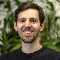 a young man with short dark hair and a light beard smiles warmly at the camera he wears a black shirt and stands against a softly blurred background filled with vibrant green plants adding a refreshing natural ambiance to the scene digital production A young man with short dark hair and a light beard smiles warmly at the camera. He wears a black shirt and stands against a softly blurred background filled with vibrant green plants, adding a refreshing, natural ambiance to the scene.