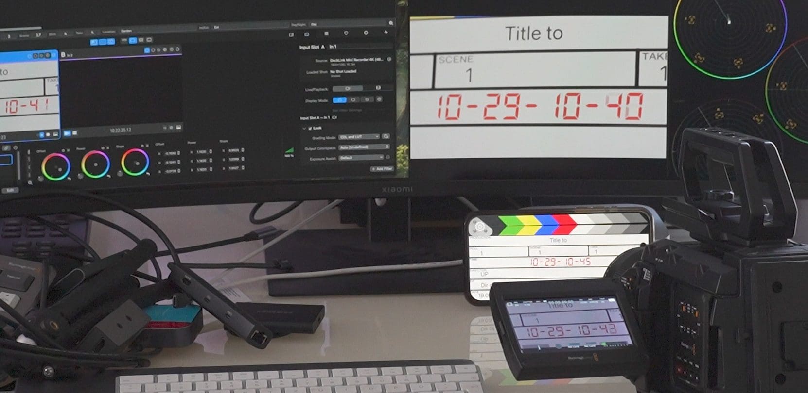 a workspace featuring two computer monitors displaying editing software and a digital slate a video camera is positioned on the right showing a timeline various cables and devices are scattered on the desk digital production A workspace featuring two computer monitors displaying editing software and a digital slate. A video camera is positioned on the right, showing a timeline. Various cables and devices are scattered on the desk.