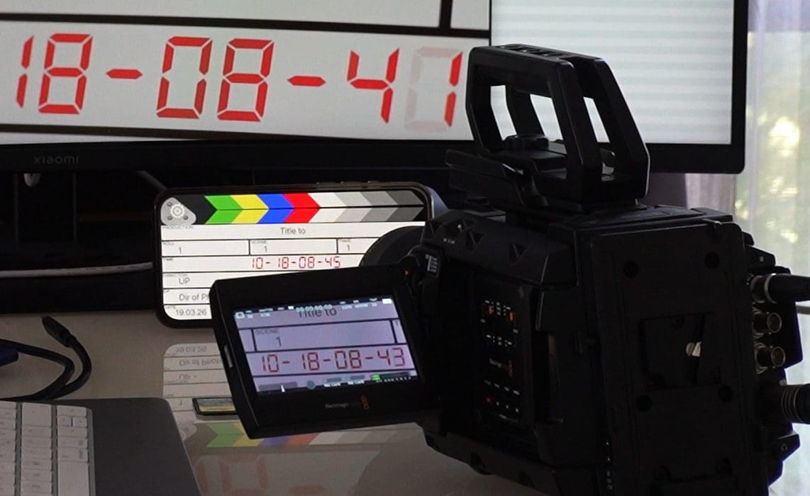 a video camera with a digital display and a clapperboard placed on a desk next to a computer with a timer displaying 180841 the camera is angled towards the desk capturing the scene digital production A video camera with a digital display and a clapperboard placed on a desk, next to a computer with a timer displaying 18:08:41. The camera is angled towards the desk, capturing the scene.