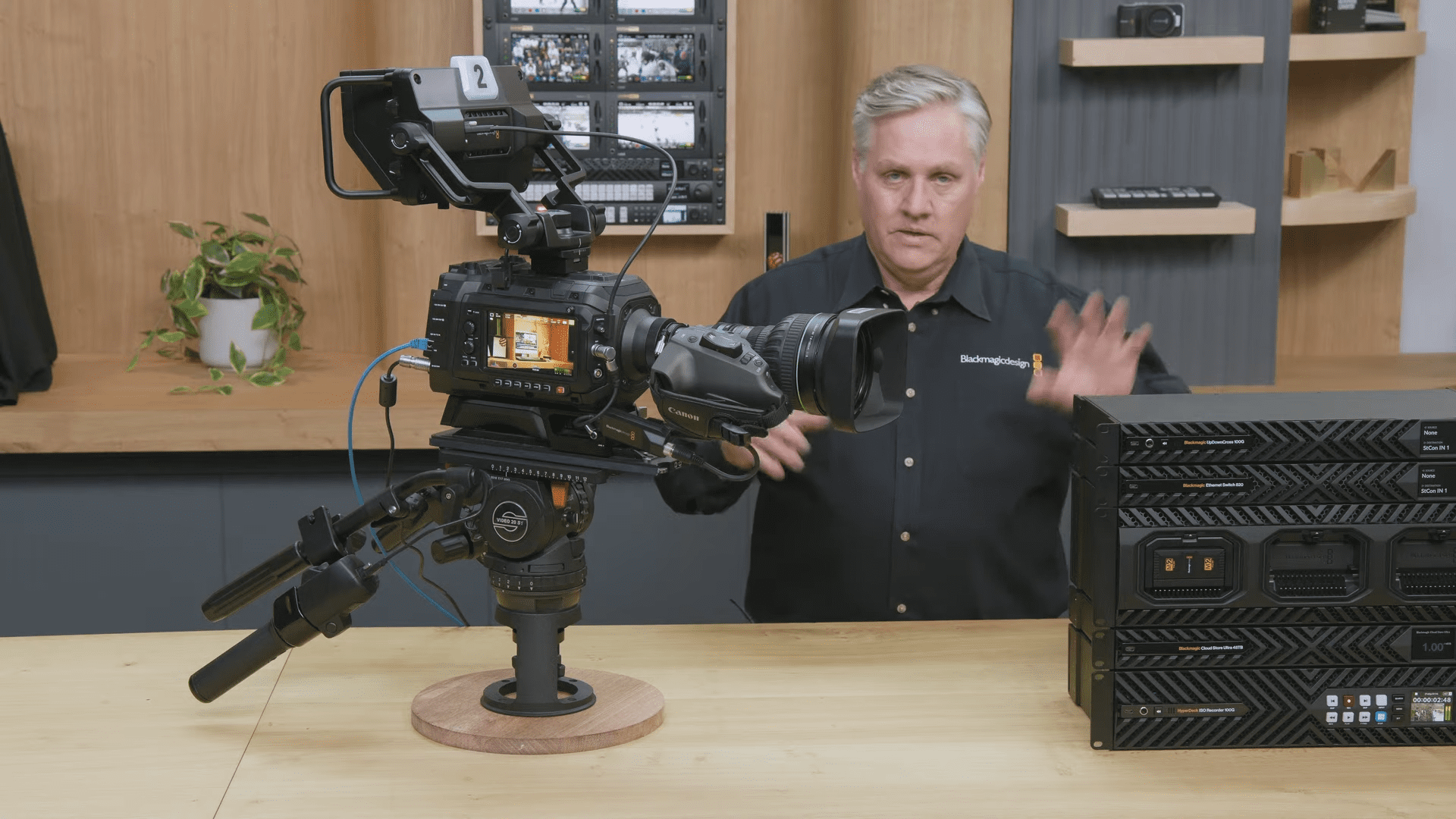 a man in a black shirt gestures with his right hand while discussing camera equipment in front of him a professional video camera is mounted on a tripod the background features shelves with additional photographic gear and a small green plant adds a touch of life to the workspace digital production A man in a black shirt gestures with his right hand while discussing camera equipment. In front of him, a professional video camera is mounted on a tripod. The background features shelves with additional photographic gear, and a small green plant adds a touch of life to the workspace.