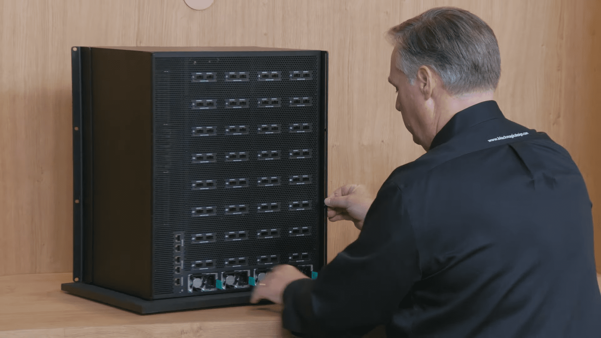 a technician in a black collared shirt is interacting with a large dark server cabinet on a wooden surface the cabinet features multiple rows of ports and connections while the technician carefully inserts a cable focusing intently on his task digital production A technician in a black collared shirt is interacting with a large, dark server cabinet on a wooden surface. The cabinet features multiple rows of ports and connections, while the technician carefully inserts a cable, focusing intently on his task.