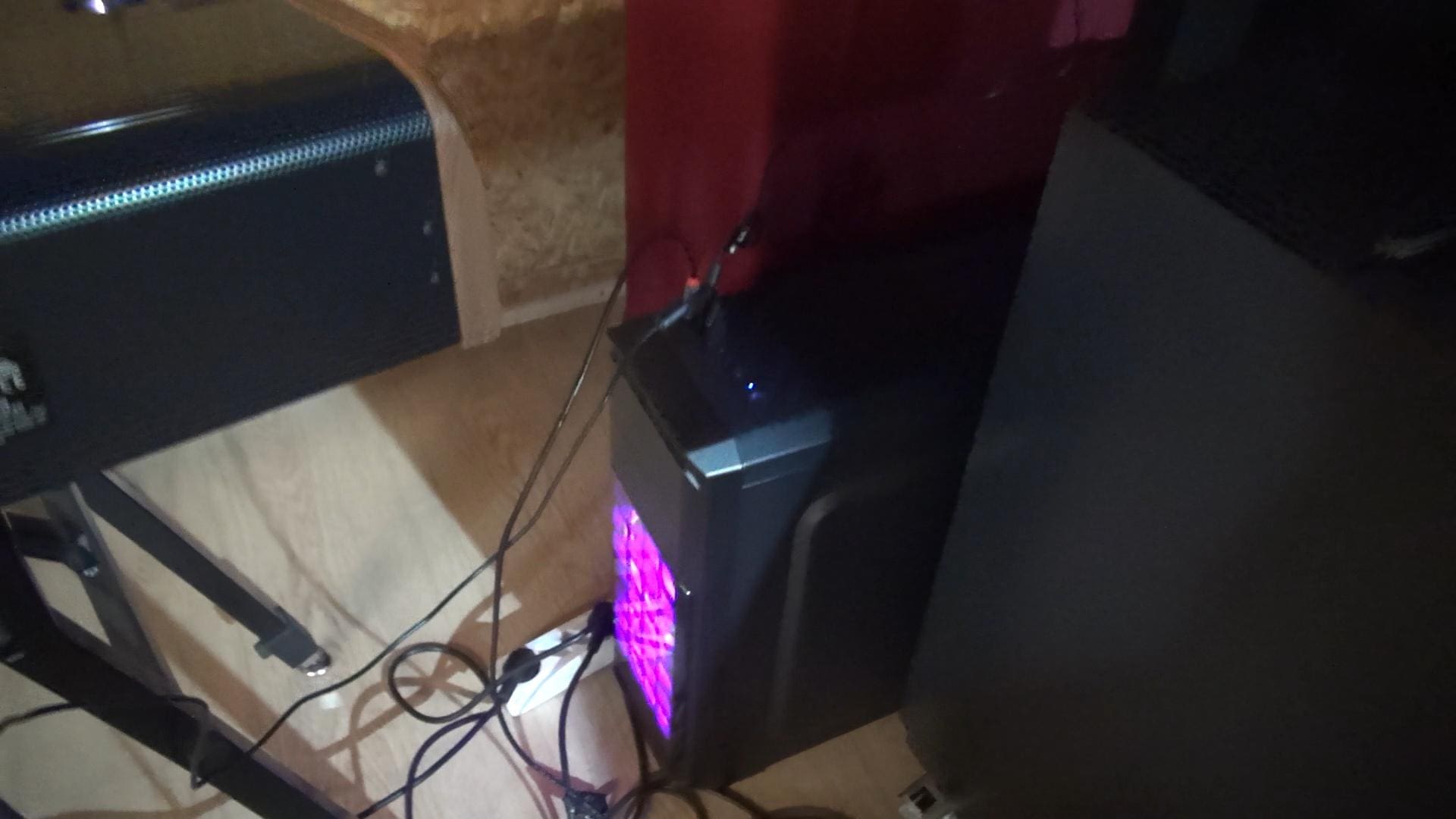 a gaming pc tower situated in a corner emitting a soft purple glow from its front fan wires are visibly tangled around the base connecting devices the floor features a wooden texture and the background includes a red wall and a black cabinet digital production A gaming PC tower situated in a corner, emitting a soft purple glow from its front fan. Wires are visibly tangled around the base, connecting devices. The floor features a wooden texture, and the background includes a red wall and a black cabinet.