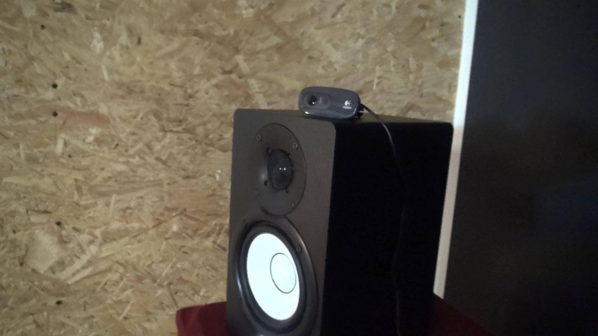 a black speaker stands on a red surface featuring a prominent white circular speaker cone atop the speaker a small dark device with a lens and a cable extends indicating it might be a camera or a microphone set against a textured wooden background digital production A black speaker stands on a red surface, featuring a prominent white circular speaker cone. Atop the speaker, a small dark device with a lens and a cable extends, indicating it might be a camera or a microphone, set against a textured wooden background.