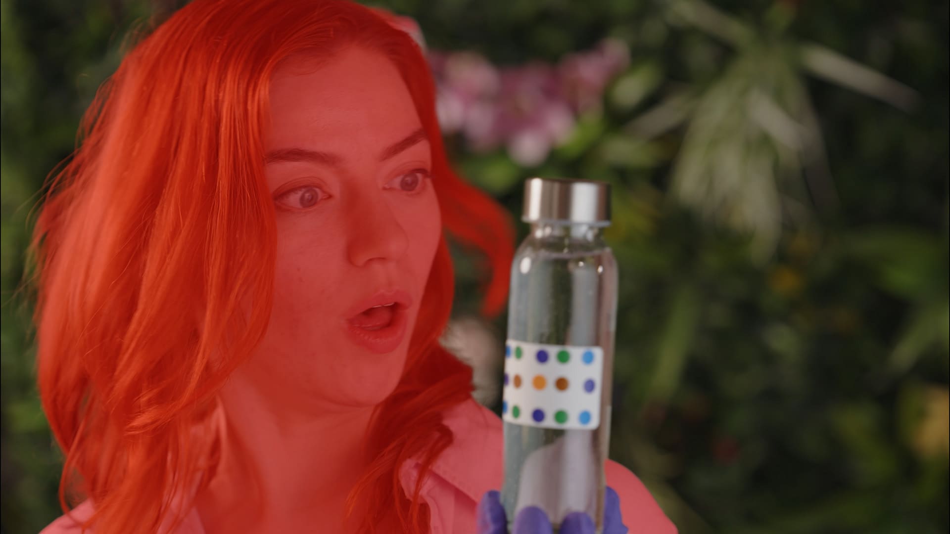 a young woman with vibrant red hair looks astonished as she gazes at a clear glass bottle featuring a whimsical label with colorful dots her surprised expression is emphasized by her bright pink shirt and the lush green backdrop filled with tropical plants digital production A young woman with vibrant red hair looks astonished as she gazes at a clear glass bottle featuring a whimsical label with colorful dots. Her surprised expression is emphasized by her bright pink shirt and the lush green backdrop filled with tropical plants.