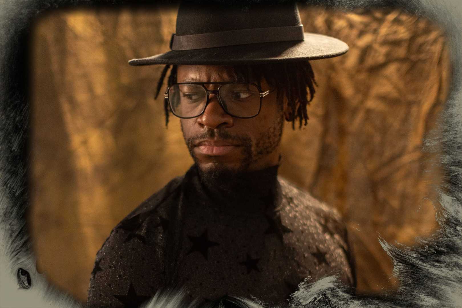 a man wearing large round glasses and a widebrimmed black hat stands against a shimmering golden backdrop his fitted longsleeved shirt is adorned with velvet black stars adding a rich texture the warm lighting highlights the intricate details of his attire creating a striking visual contrast digital production A man wearing large round glasses and a wide-brimmed black hat stands against a shimmering golden backdrop. His fitted, long-sleeved shirt is adorned with velvet black stars, adding a rich texture. The warm lighting highlights the intricate details of his attire, creating a striking visual contrast.