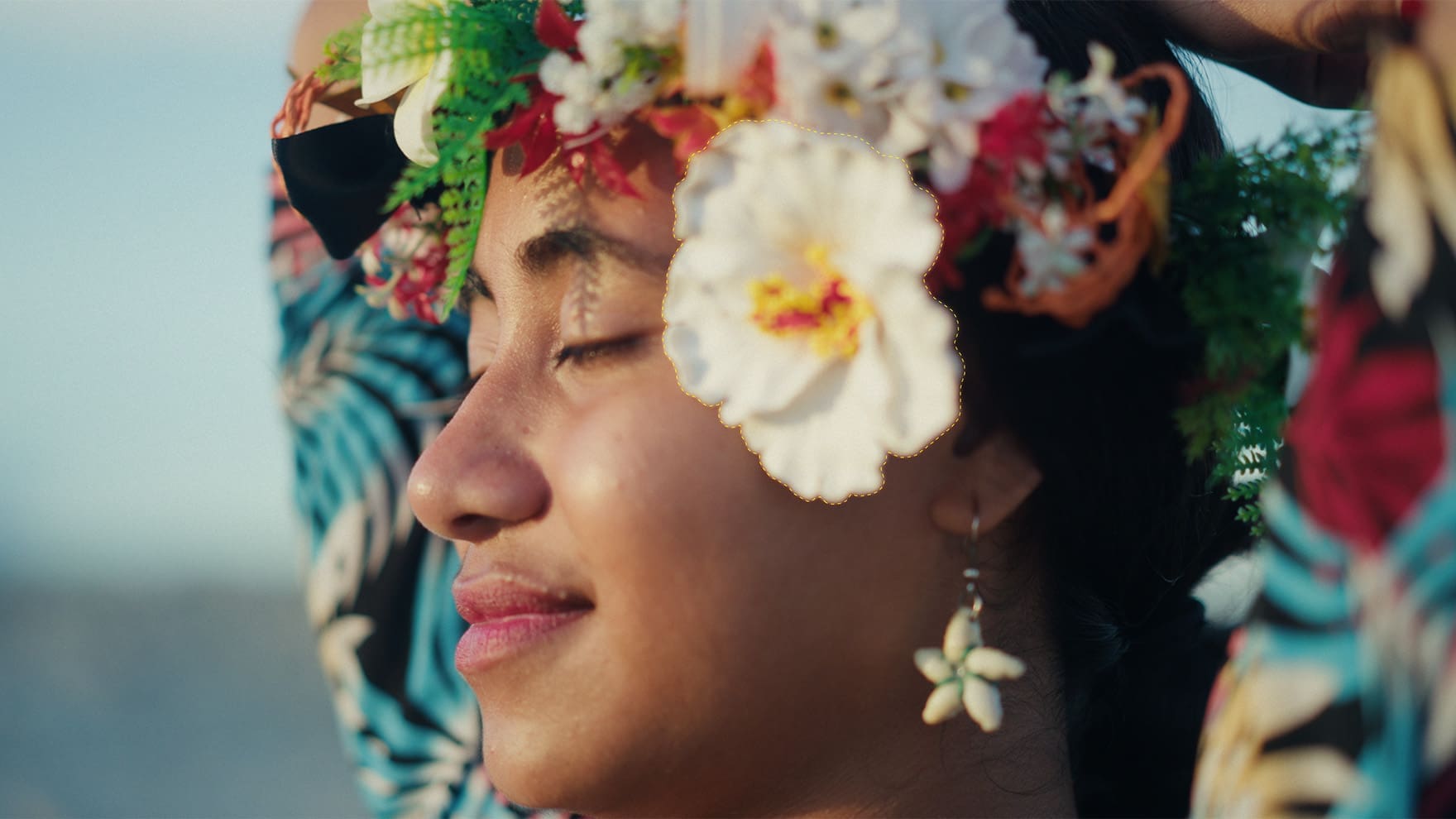 mochapro20265matteassistmlsearcharea digital production Close-up of a woman with a colorful flower crown and hibiscus earrings, eyes closed on a sunny day at the beach.