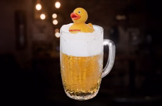 A clear beer mug filled with golden lager sits against a dark, softly blurred background, topped with a white foam cap and a small yellow rubber duck labeled MAXON. Warm highlights catch the glass handle and bubbles, giving the composition a playful promotional feel.