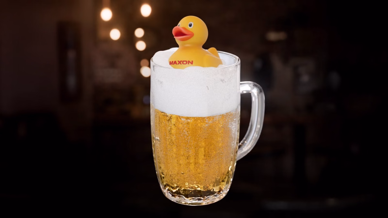 A clear beer mug filled with golden lager sits against a dark, softly blurred background, topped with a white foam cap and a small yellow rubber duck labeled MAXON. Warm highlights catch the glass handle and bubbles, giving the composition a playful promotional feel.