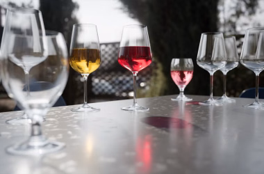 A row of wine glasses on a glossy outdoor table, including clear, amber, and pink-filled stems arranged across the foreground and midground. Soft daylight reflects off the glass and smooth surface, with a blurred dark background that gives the scene a calm, elegant look.